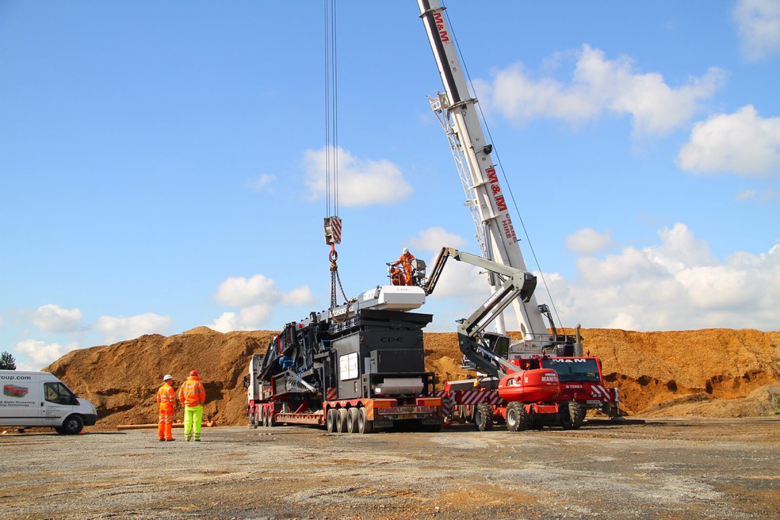 A mobile crane lifting a large machine off a low-loader lorry on an open site, with two workers in hi-vis watching