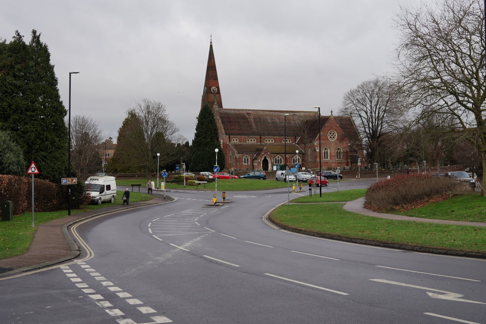 A red-brick church with a tall spire beyond a roundabout and grass verges in Burgess Hill