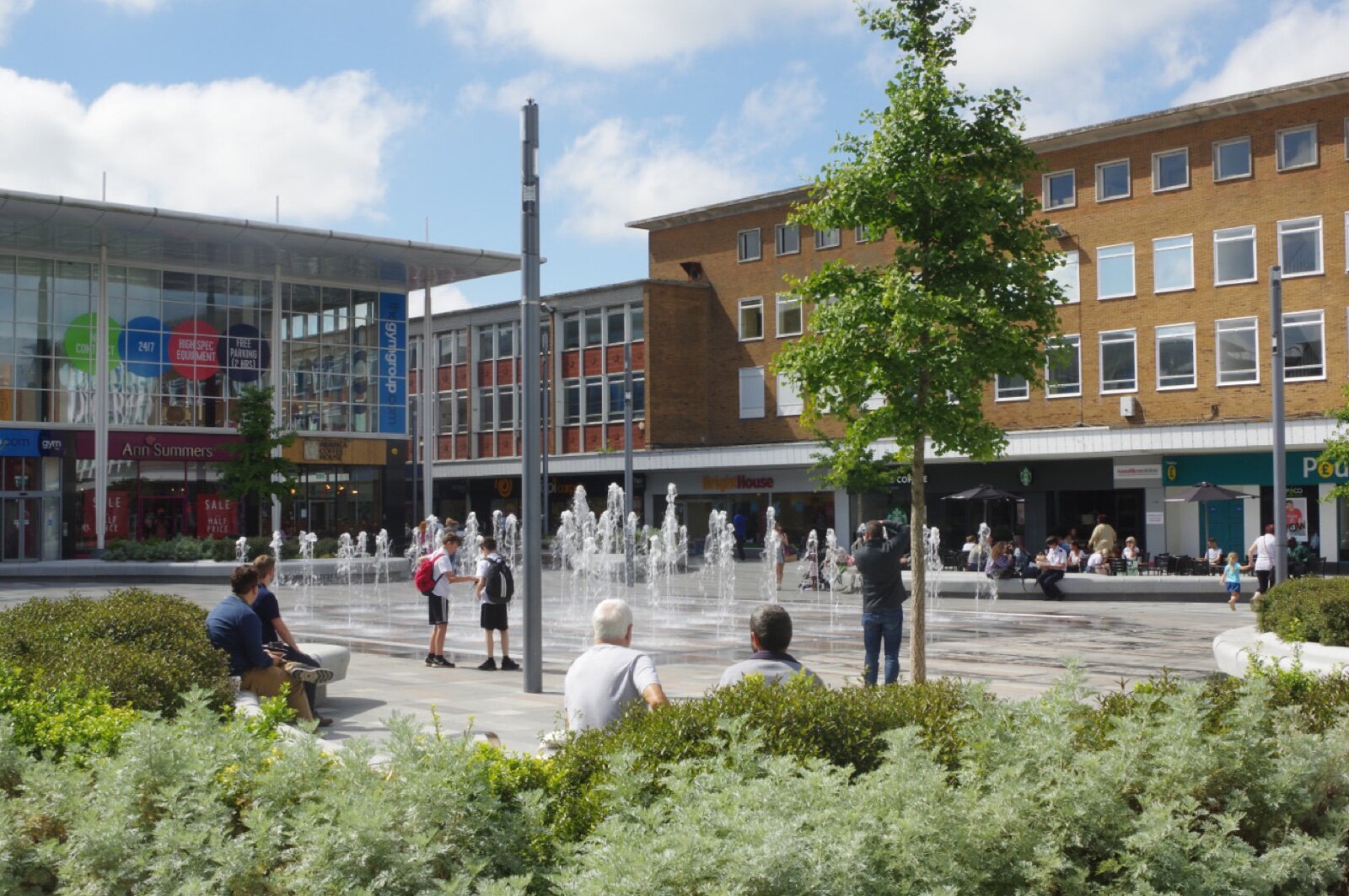 Queens Square in Crawley town centre with fountains set into the paving, young trees, planting and shopfronts under a blue sky