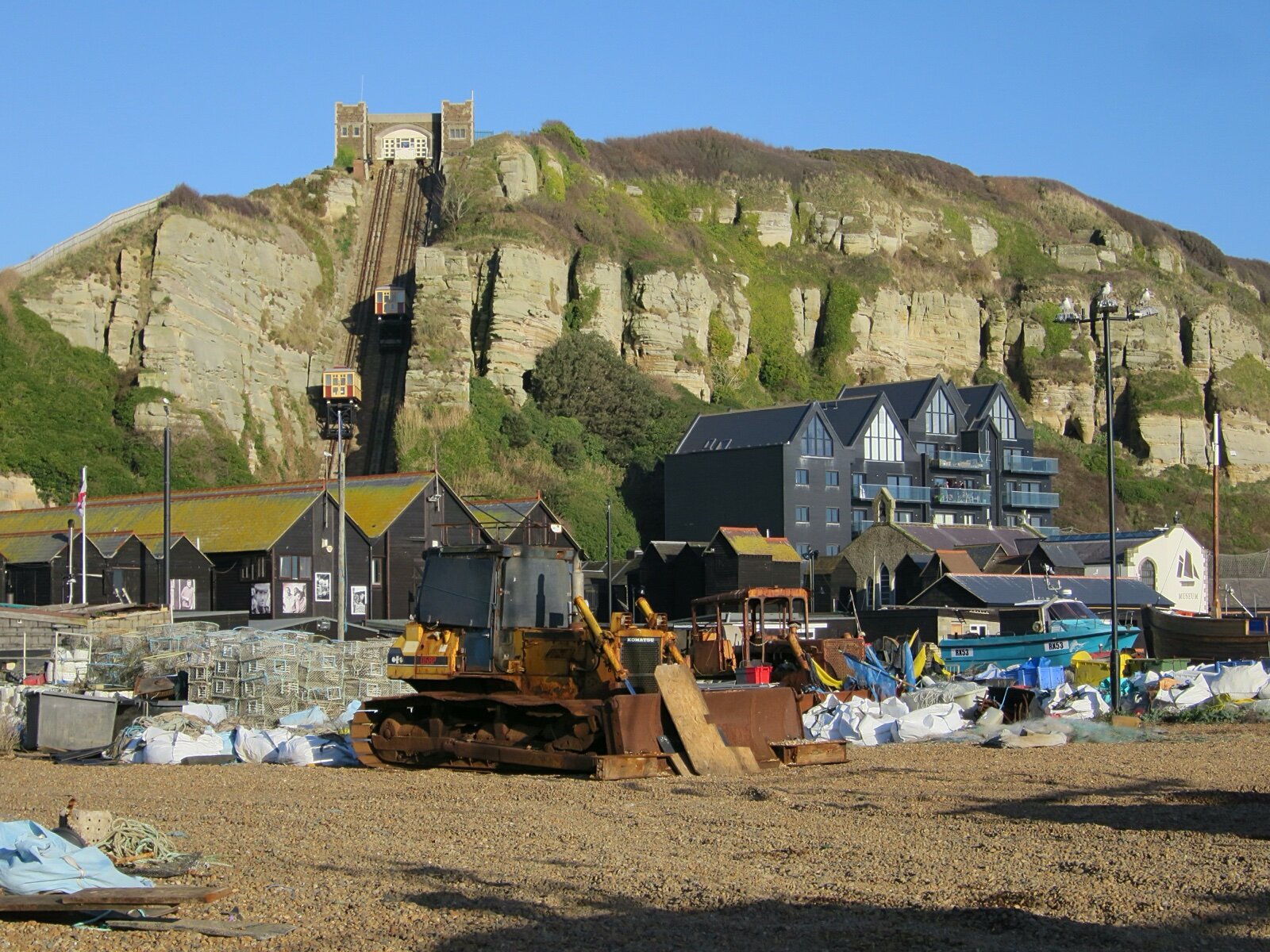 The Stade at Hastings Old Town with tall black net huts and fishing gear on the shingle, below the sandstone cliff of East Hill and its cliff railway